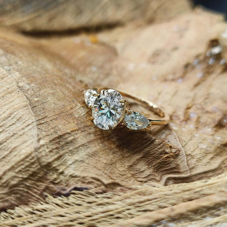May include: A gold ring with an oval-cut center stone and two pear-shaped side stones. The ring is set on a gold band and is displayed on a wooden surface. The stones sparkle brightly.