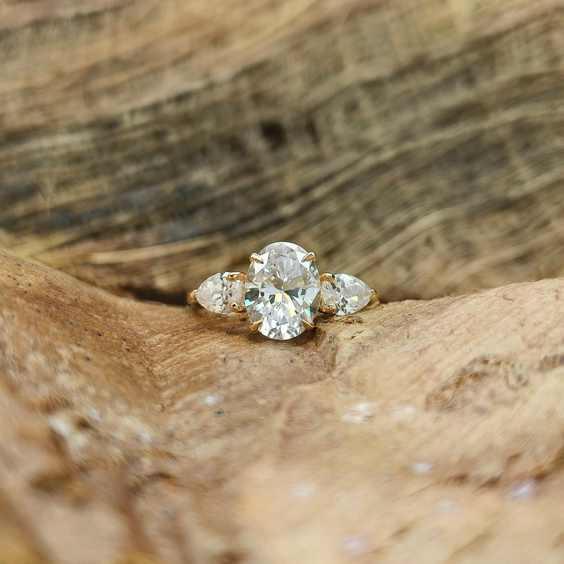 May include: A gold ring featuring a large oval-cut diamond center stone, flanked by two pear-shaped diamonds. The ring is set on a gold band, showcasing a classic three-stone design. The ring is displayed on a wooden surface.