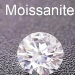 May include: A round, brilliant-cut moissanite gemstone. The gemstone is clear and faceted, reflecting light. The word "Moissanite" is written in white text above the gemstone against a dark gray textured background.