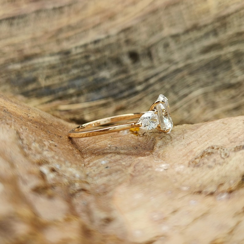 May include: A gold ring with a large, rectangular, clear gemstone and two smaller pear-shaped gemstones. The ring is set on a wooden surface, showcasing the ring's design and sparkle.