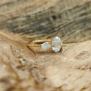 May include: A gold engagement ring with a large oval-cut center stone and two pear-shaped side stones. The ring is set on a gold band and is displayed on a wooden surface.