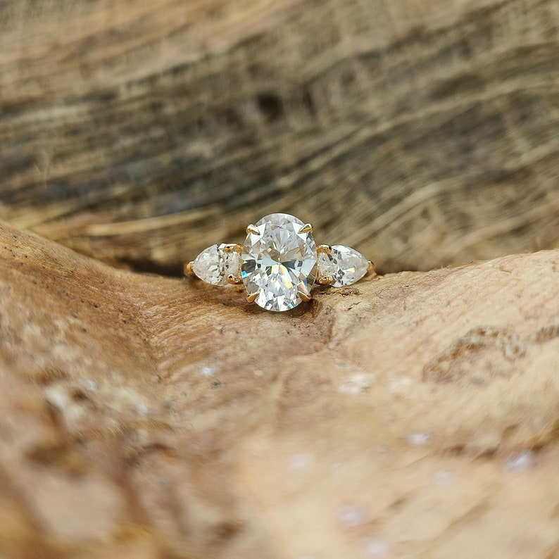 May include: A gold engagement ring with a large oval-cut center stone and two pear-shaped side stones. The ring is set on a wooden surface, showcasing the ring's sparkle and design. The ring's band is a simple, elegant design.