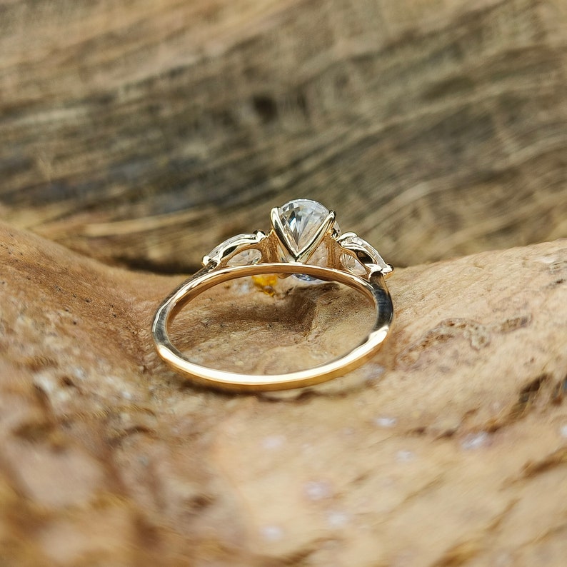 May include: A gold ring with a large, pear-shaped, clear gemstone as the centerpiece. Two smaller, clear gemstones flank the main stone. The ring is set on a wooden surface, showcasing the ring's design and sparkle.