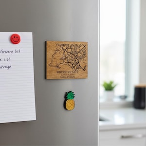 Puede incluir: Un imán cuadrado de madera con un diseño de mapa y el texto "WHERE WE MET". Un imán de piña está debajo. Un imán de cara sonriente roja y una lista de compras también están en el refrigerador.