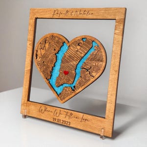 Puede incluir: Arte de mapa en forma de corazón de madera con un río azul y un corazón rojo que marca una ubicación. El mapa está enmarcado en un marco de madera marrón claro. El texto "Rafael & Natalia" y "Where We Fell in Love 19.01.2023" están grabados.