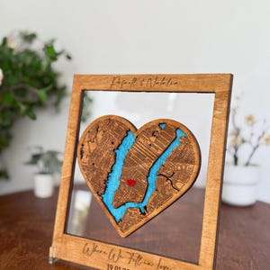 Könnte beinhalten: Holzgerahmte herzförmige Karte von New York City mit einem roten Herz, das einen Ort markiert. Die Karte ist vor blauem Hintergrund, mit dem Text "Where We Fell in Love" und einem Datum. Der Rahmen ist aus hellbraunem Holz.
