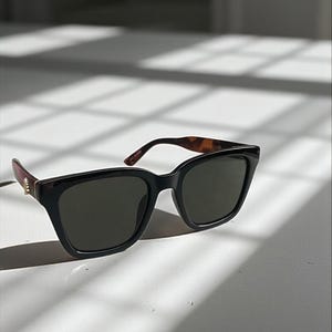 May include: Black sunglasses with dark lenses and a thick, square frame. The temples have a tortoiseshell pattern and the arms are a dark brown colour. The sunglasses are on a white surface, with shadows from a window.