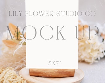 May include: A white card mockup with the text "LILY FLOWER STUDIO CO MOCK UP 5X7" on a wooden stand. The card is surrounded by dried flowers and grasses on a light beige surface.
