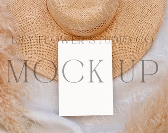 May include: A white card mockup with the word "MOCKUP" printed in gray text. The card is lying on a light beige fabric surface with a straw hat and dried pampas grass.