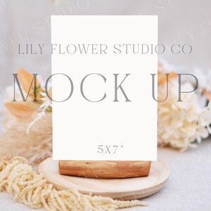 Może przedstawiać: Biała karta mockupowa z tekstem "LILY FLOWER STUDIO CO MOCK UP 5X7" na drewnianym stojaku. Karta jest otoczona suszonymi kwiatami i trawami na jasnobeżowej powierzchni.