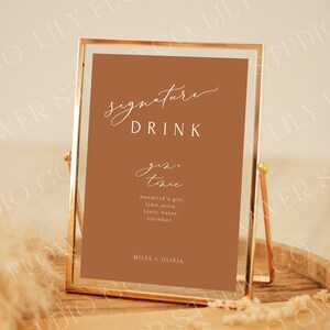 Op de afbeelding: Een gouden lijst met een bruine achtergrond en witte tekst die "signature drink gin + tonic hendrick's gin, lime juice, tonic water, cucumber MILES + OLIVIA" luidt.