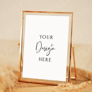 Op de afbeelding: Een gouden metalen fotolijst met een witte achtergrond en de tekst "YOUR Design HERE" in zwart. De lijst staat op een houten dienblad met een beige, pluche textuur.
