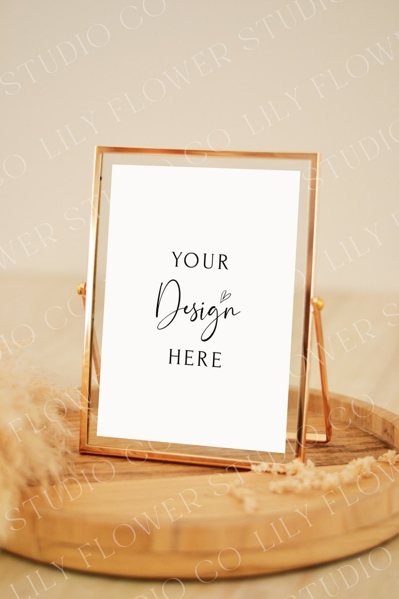 Op de afbeelding: Een gouden metalen fotolijst met een witte achtergrond en de tekst "YOUR Design HERE" in zwart. De lijst staat op een houten dienblad met een beige, pluche textuur.