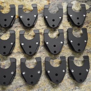 Puede incluir: Doce abrazaderas de plástico negro con tornillos plateados. Las abrazaderas tienen un diseño en forma de U y están dispuestas en tres filas de cuatro.