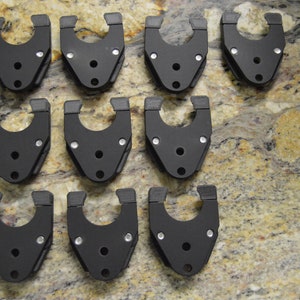 May include: Twelve black plastic clips with a U-shaped design and multiple silver screws. The clips are arranged in three rows of four on a mottled brown and gold surface.