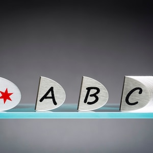 May include: Four silver metal tags with a red star and the letters A, B, and C. The tags are arranged in a row on a glass surface.