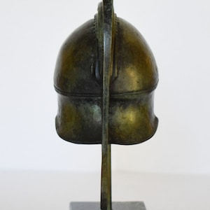 May include: A bronze sculpture of a Greek helmet on a black marble base. The helmet has a curved crest and is detailed with intricate carvings.