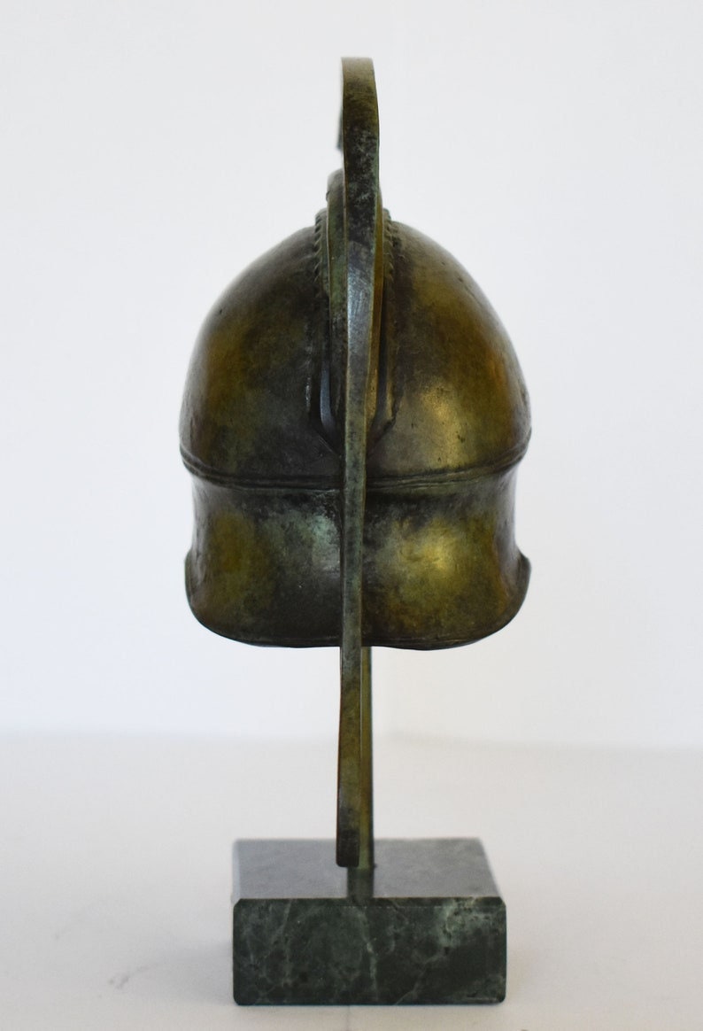 May include: A bronze sculpture of a Greek helmet on a black marble base. The helmet has a curved crest and is detailed with intricate carvings.