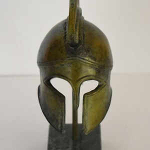 May include: A bronze Greek warrior helmet with a plume on top, displayed on a black marble base.