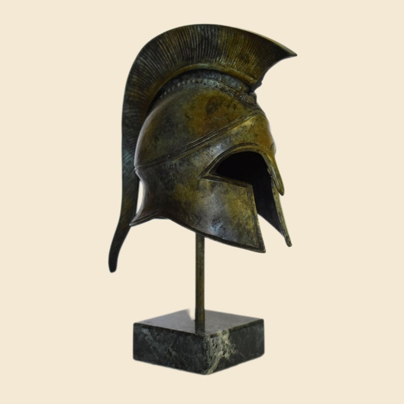 May include: A bronze replica of a Greek warrior helmet with a plume on top, displayed on a black marble base.