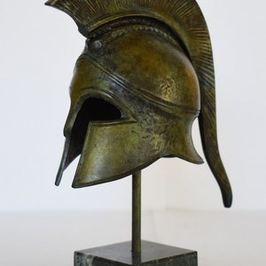 May include: A bronze Greek warrior helmet with a plume on top, displayed on a marble base.