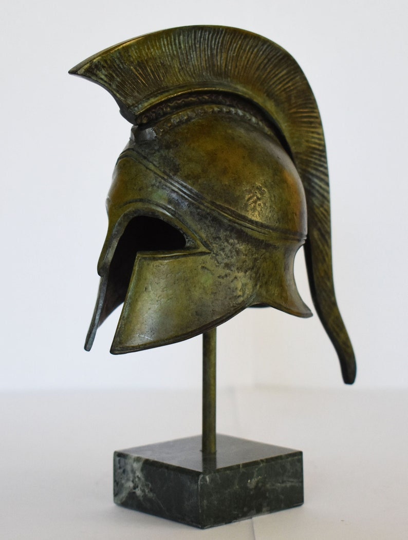 May include: A bronze Greek warrior helmet with a plume on top, displayed on a marble base.