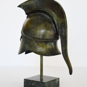 May include: A bronze replica of a Greek warrior helmet with a plume on top, displayed on a black marble base.