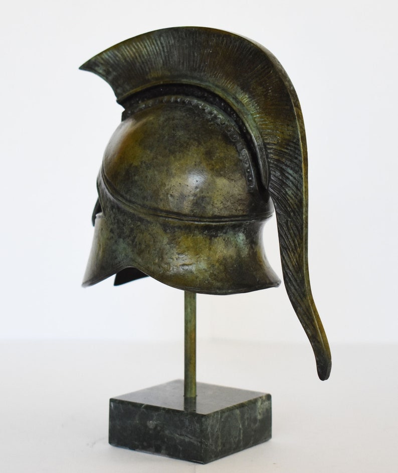 May include: A bronze replica of a Greek warrior helmet with a plume on top, displayed on a black marble base.