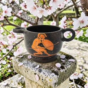 May include: A black ceramic cup with two handles featuring an orange-toned illustration of a crouching warrior wearing a helmet and holding a round shield and a spear. The vessel has a wide, rounded body and a sturdy base, showcasing a classic design.