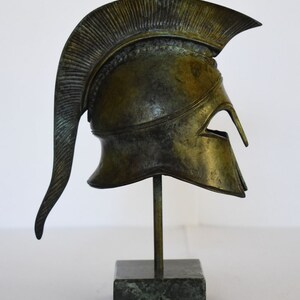 May include: A bronze replica of a Greek warrior helmet with a plume on top, displayed on a green marble base.