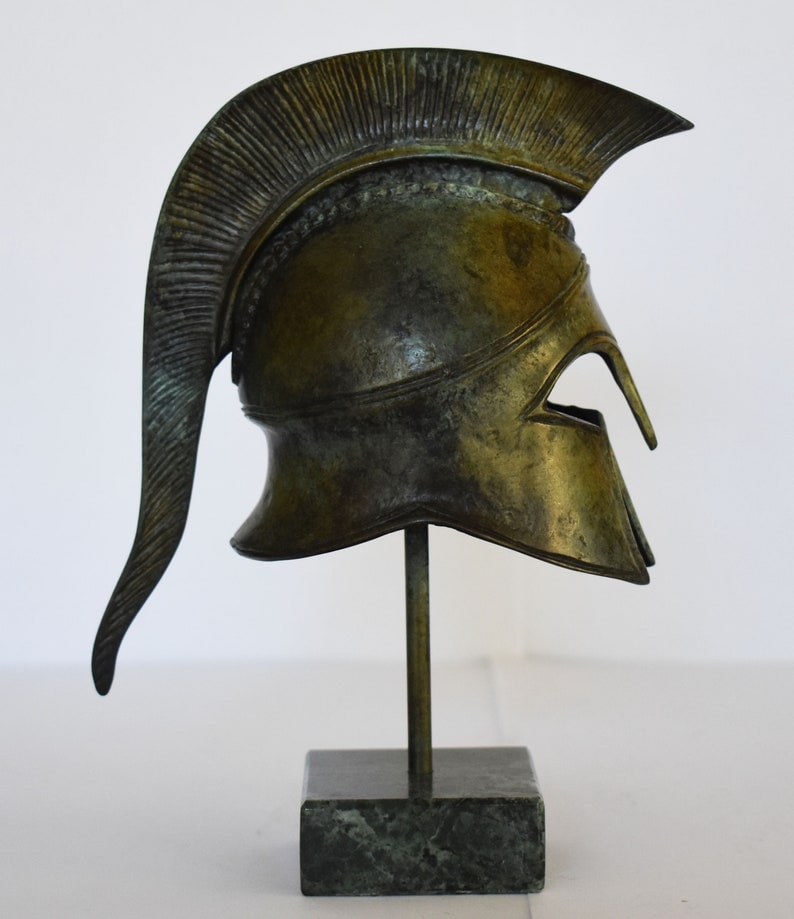 May include: A bronze replica of a Greek warrior helmet with a plume on top, displayed on a green marble base.