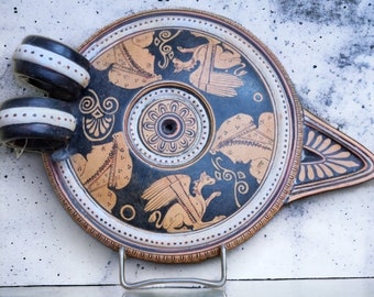 May include: A black and white ceramic oil lamp with a round base and a spout. The lamp is decorated with a stylized design of winged creatures and floral motifs.