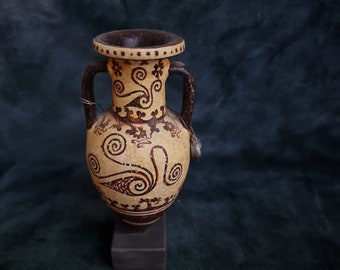 May include: A brown and beige ceramic amphora with a geometric design. The amphora has two handles and a narrow neck. It is sitting on a dark brown base.