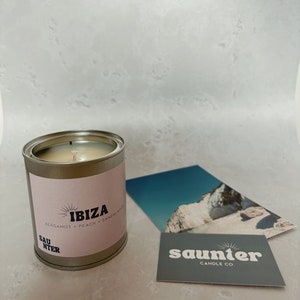 Op de afbeelding: Een zilveren kaarsenblik met een roze etiket waarop "IBIZA" en "BERGAMOT + PEACH + SANDALWOOD" staat. Het etiket heeft ook de woorden "SAUNTER" en "CANDLE CO.". De kaars staat op een wit oppervlak met een blauw-witte ansichtkaart en een grijze visitekaart met de woorden "SAUNTER CANDLE CO.".