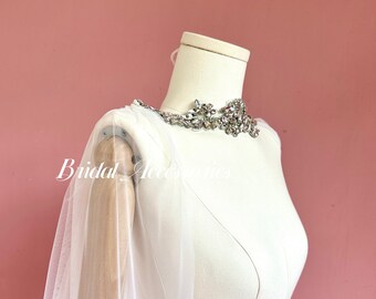 Puede incluir: Una capa de novia de tul blanco con un adorno de cristal en el escote. La capa está drapeada sobre un maniquí blanco.