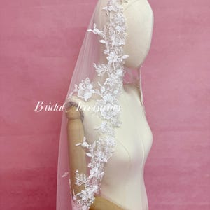 May include: A white bridal veil featuring intricate floral lace embroidery along the edges, adorned with small clear beads and sequins. The veil is draped over a mannequin, and the text Bridal Accessories is visible on the left side of the image.