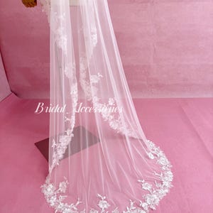 May include: A long, sheer white veil featuring intricate white floral lace embroidery along the edges. The delicate lace pattern extends from the top down to the rounded hem. The text Bridal Accessories is printed in white on the left side.