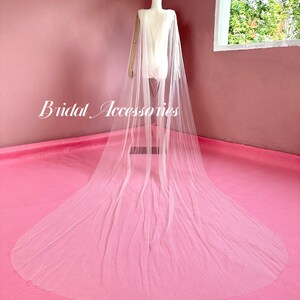 Pu&ograve; includere: Un velo da sposa in tulle bianco con un lungo strascico, drappeggiato su un manichino su sfondo rosa. Il velo &egrave; trasparente e delicato, con un design semplice. Il testo "Bridal Accessories" &egrave; visibile nell'immagine.
