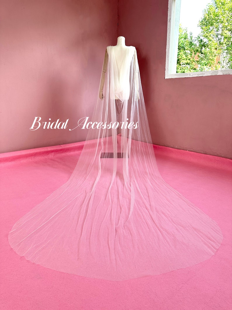 Pu&ograve; includere: Un velo da sposa in tulle bianco con un lungo strascico, drappeggiato su un manichino su sfondo rosa. Il velo &egrave; trasparente e delicato, con un design semplice. Il testo "Bridal Accessories" &egrave; visibile nell'immagine.