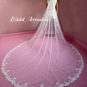 May include: A long white cathedral-length bridal veil featuring intricate white floral lace embroidery along the edges and a sheer tulle fabric. The text "Bridal Accessories" is printed in white script on the left side of the veil.