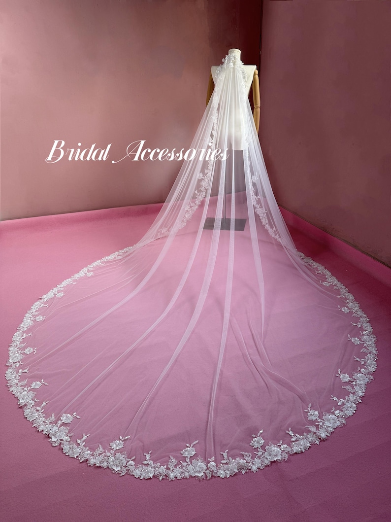 May include: A long white cathedral-length bridal veil featuring intricate white floral lace embroidery along the edges and a sheer tulle fabric. The text "Bridal Accessories" is printed in white script on the left side of the veil.