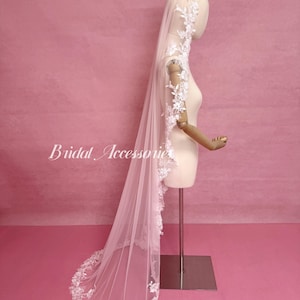 May include: A long, sheer, light pink bridal veil featuring intricate white floral lace embroidery along the edges. The veil is draped over a mannequin, with the lace detailing extending from the head down to the floor-length hem.