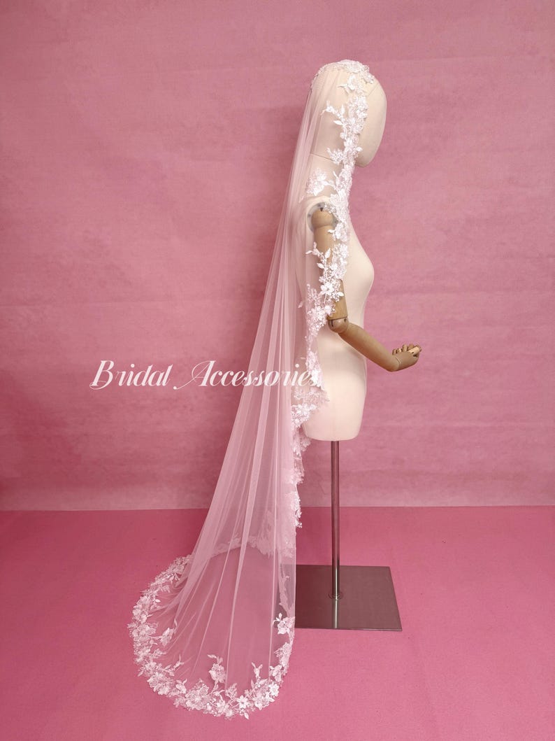 May include: A long, sheer, light pink bridal veil featuring intricate white floral lace embroidery along the edges. The veil is draped over a mannequin, with the lace detailing extending from the head down to the floor-length hem.
