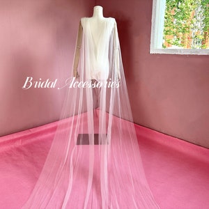Pu&ograve; includere: Un'ampia cappa da sposa in tulle bianco con una lunga coda. La cappa &egrave; drappeggiata su un manichino ed &egrave; esposta su una superficie rosa.