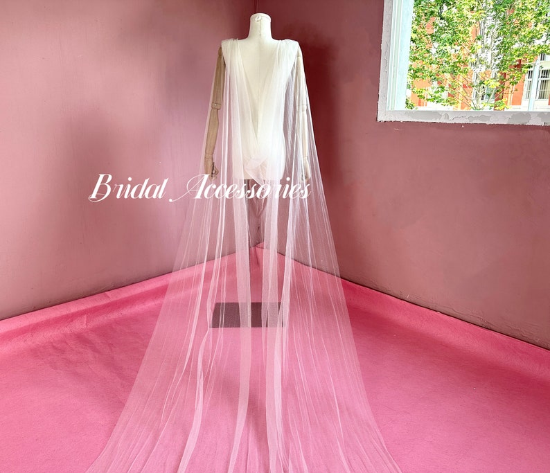 Pu&ograve; includere: Un'ampia cappa da sposa in tulle bianco con una lunga coda. La cappa &egrave; drappeggiata su un manichino ed &egrave; esposta su una superficie rosa.