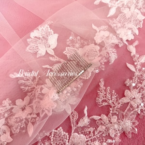 May include: A sheer light pink veil featuring white floral lace embroidery with sequins and beads along the edges. A silver-toned metal hair comb is attached to the fabric. The text Bridal Accessories is printed in white script on the veil.