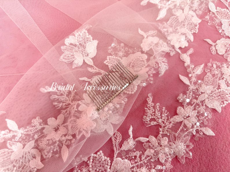 May include: A sheer light pink veil featuring white floral lace embroidery with sequins and beads along the edges. A silver-toned metal hair comb is attached to the fabric. The text Bridal Accessories is printed in white script on the veil.