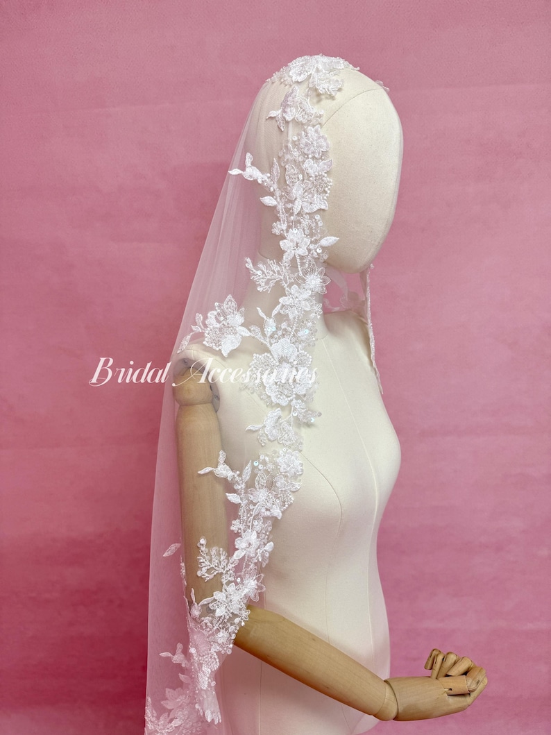 May include: A white bridal veil featuring intricate floral lace embroidery along the edges, adorned with small clear beads and sequins. The veil is draped over a mannequin, and the text Bridal Accessories is visible on the left side of the image.