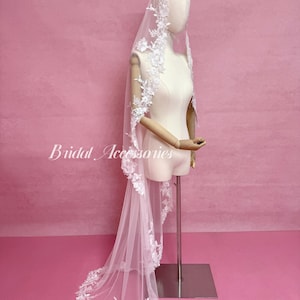 May include: A long white bridal veil featuring intricate floral lace embroidery along the edges, draped over a mannequin. The veil is made of sheer tulle fabric and displays the text Bridal Accessories in white script on the left side.