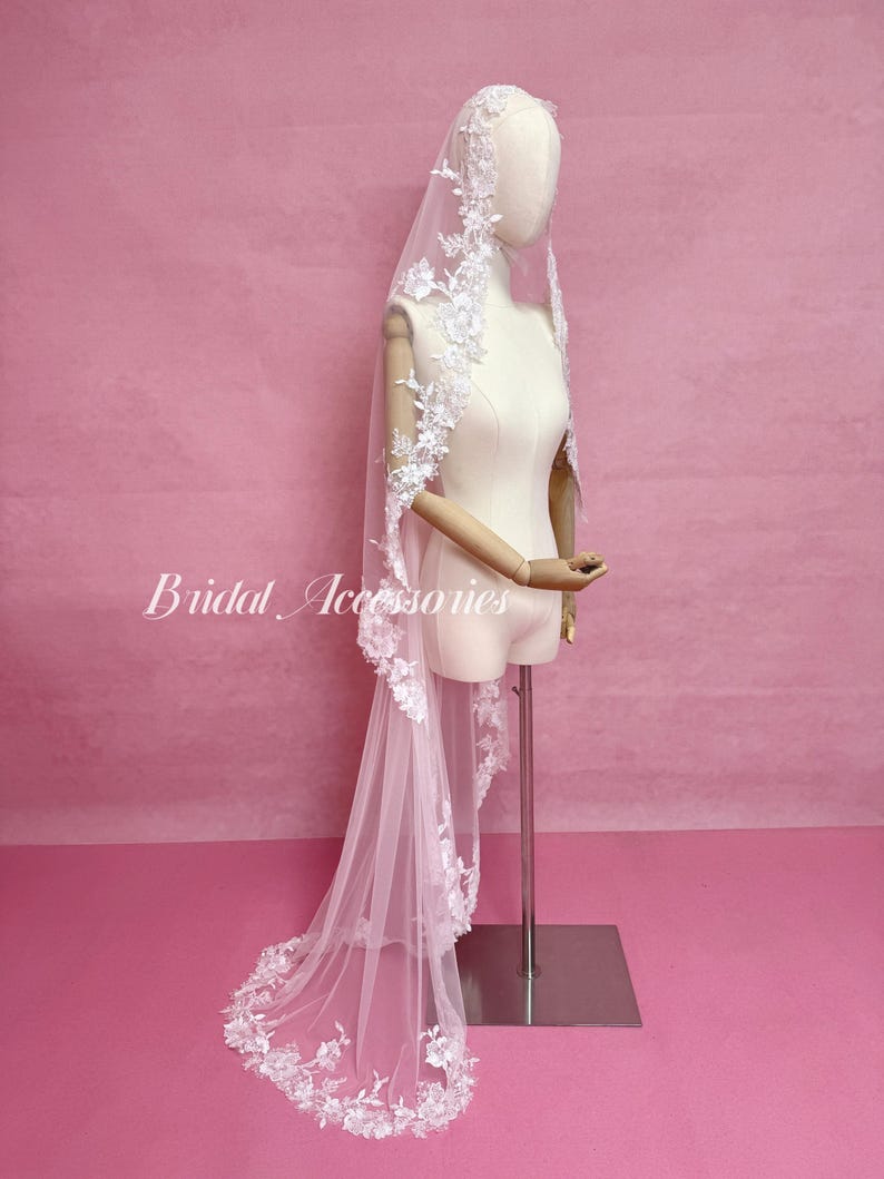 May include: A long white bridal veil featuring intricate floral lace embroidery along the edges, draped over a mannequin. The veil is made of sheer tulle fabric and displays the text Bridal Accessories in white script on the left side.